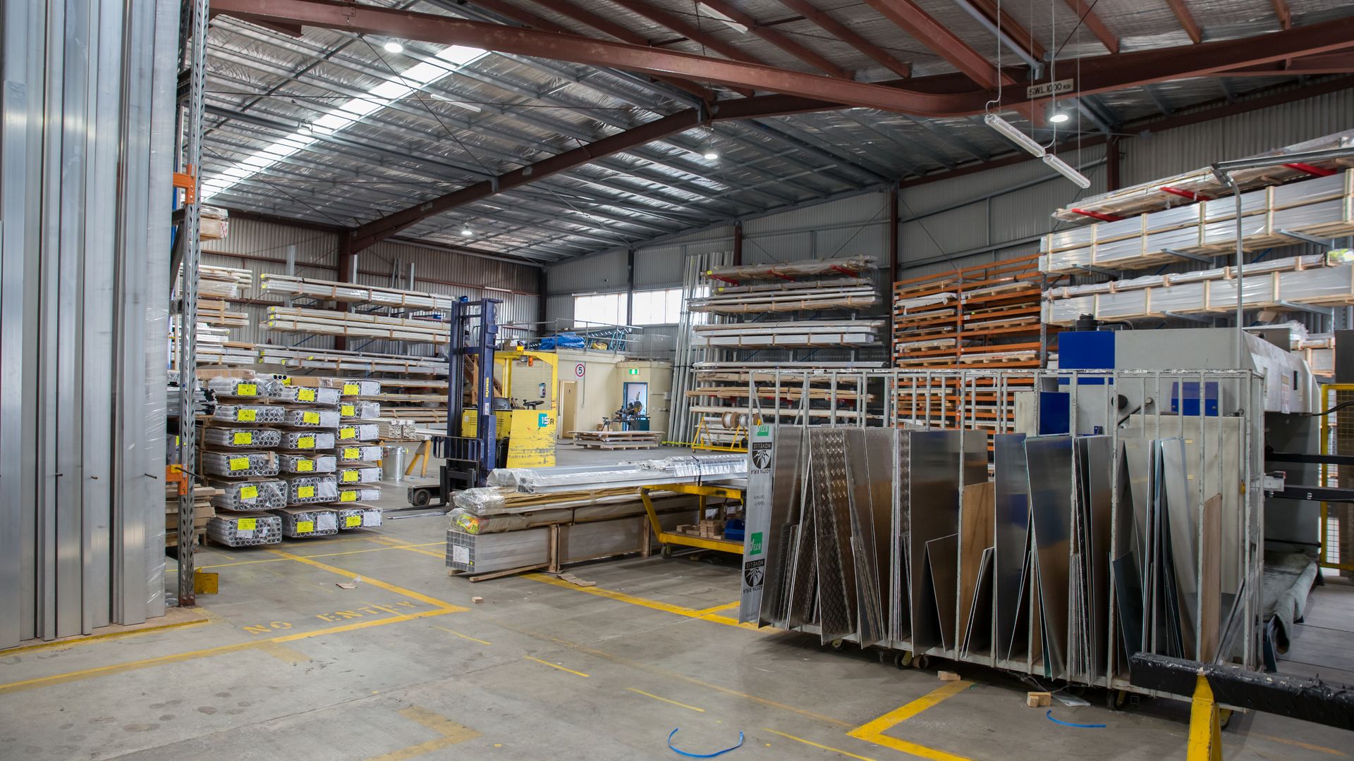 Nave de un taller de aluminio con estanterías llenas de perfiles y barras apiladas, planchas de aluminio en carros verticales y una carretilla elevadora al fondo.