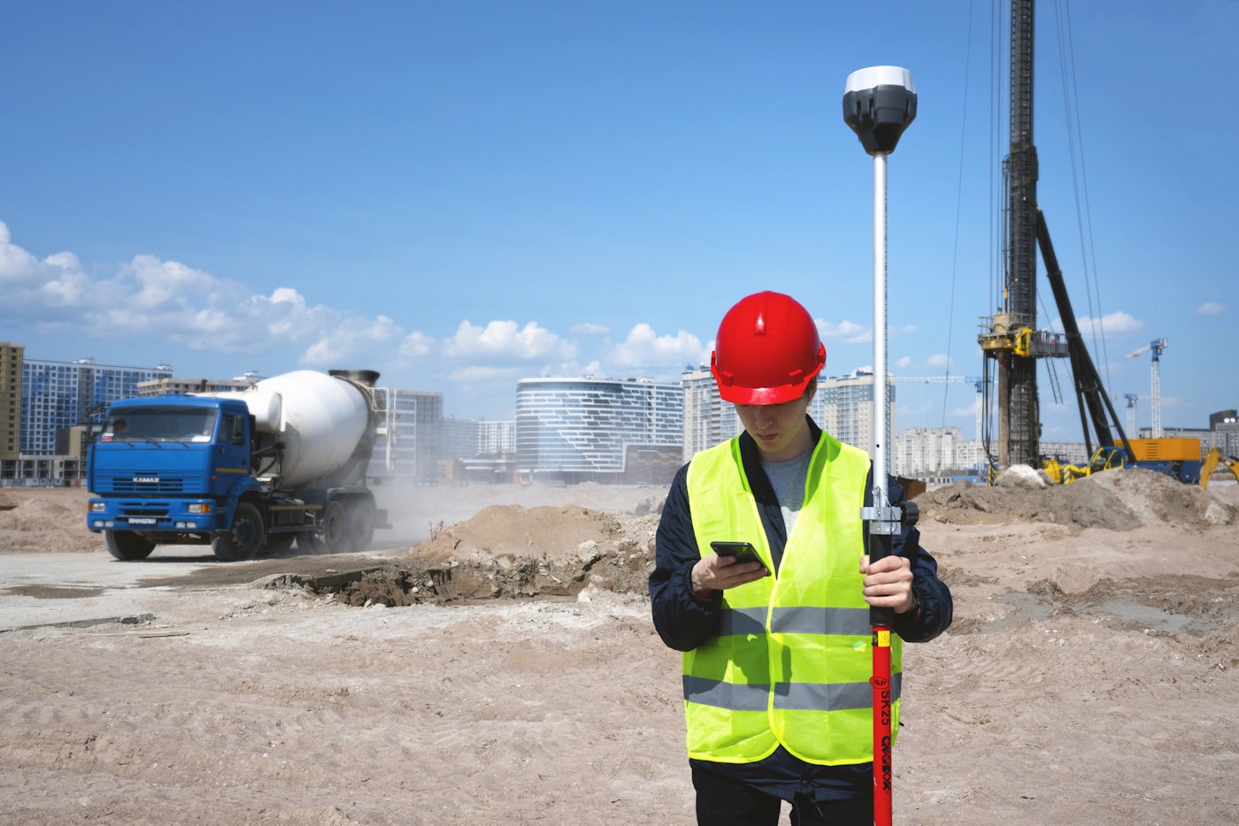 Trabajador con casco rojo y chaleco reflectante consultando su teléfono móvil en plena obra, con un camión hormigonera y una máquina de pilotaje al fondo.