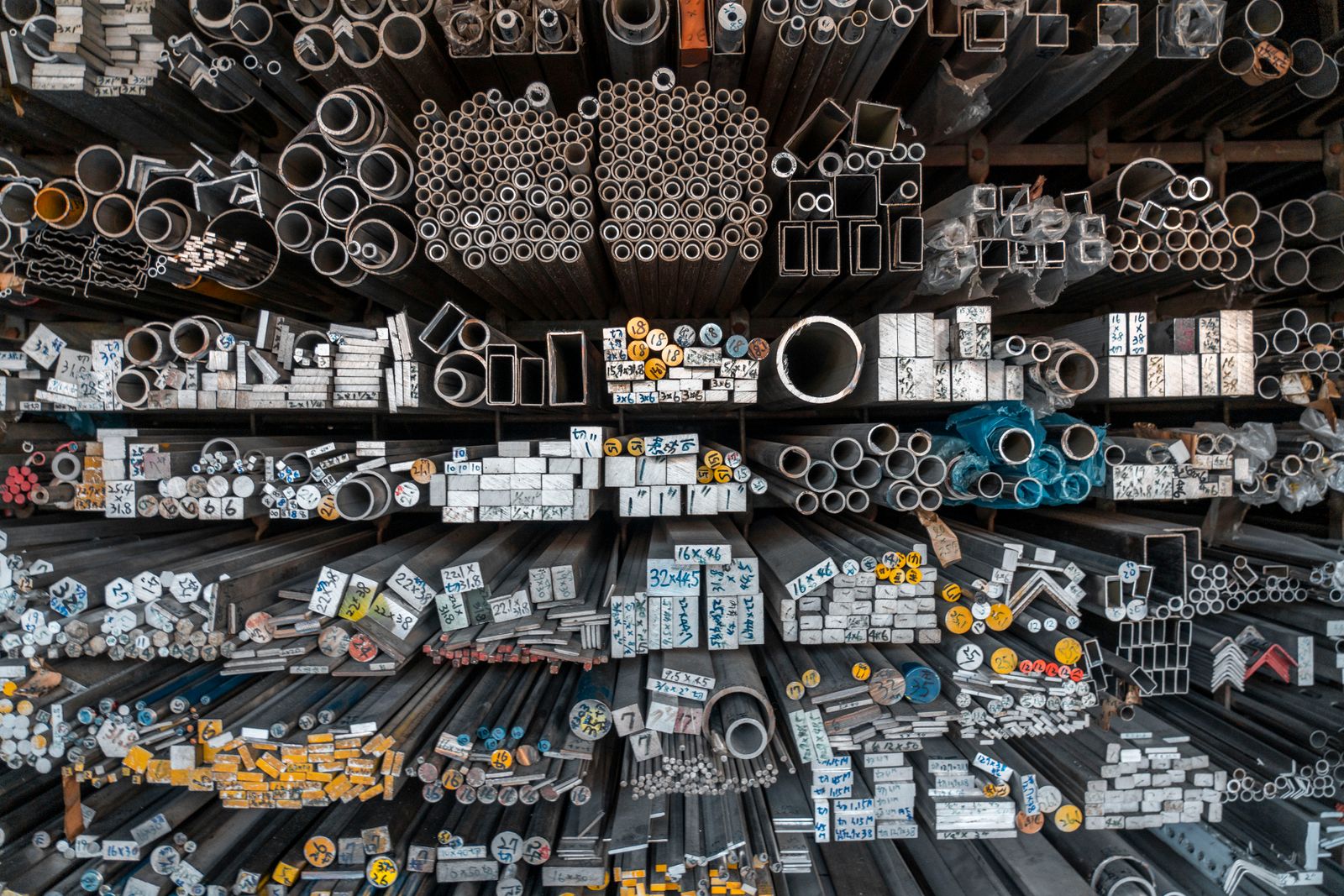 Almacén de perfilería visto de frente: estanterías con tubos redondos, tubos cuadrados, pletinas y angulares de aluminio apilados por su sección, cada montón etiquetado con su medida.