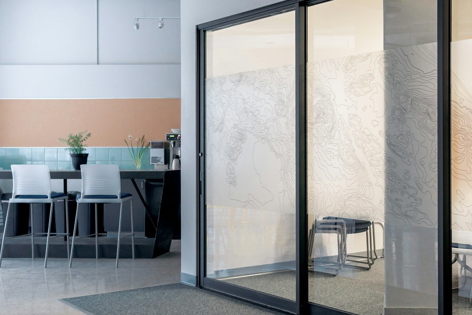 Puerta corrediza de vidrio con marco de aluminio negro instalada en un interior luminoso, junto a una zona de mesas altas y taburetes.